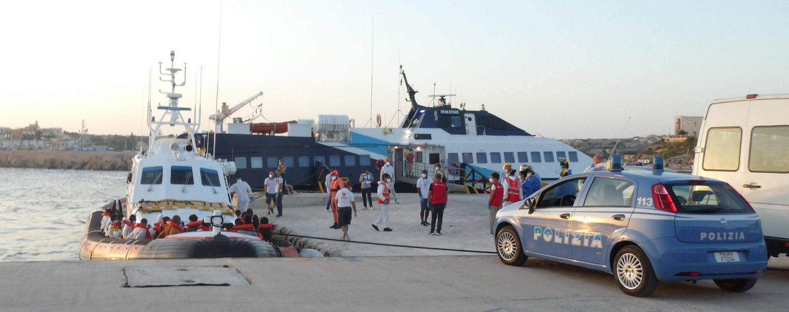 auto della polizia su una banchina con persone e soccorritori e diverse barche con persone con giubbotti di salvataggio