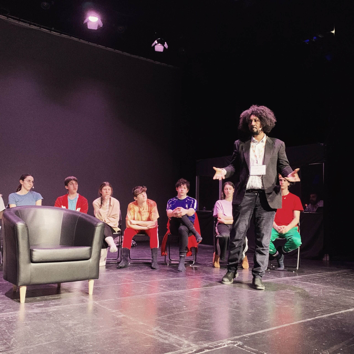 foto di un uomo su un palco con sette persone su sedie da palco che guardano