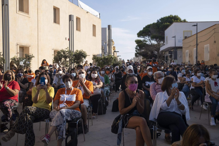 Un pubblico di oltre 100 persone su sedie che ascoltano e applaudono in una piazza fuori