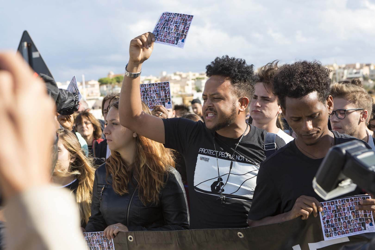 un gruppo di attivisti con un pezzo di carta in mano che mostra i volti dei migranti morti