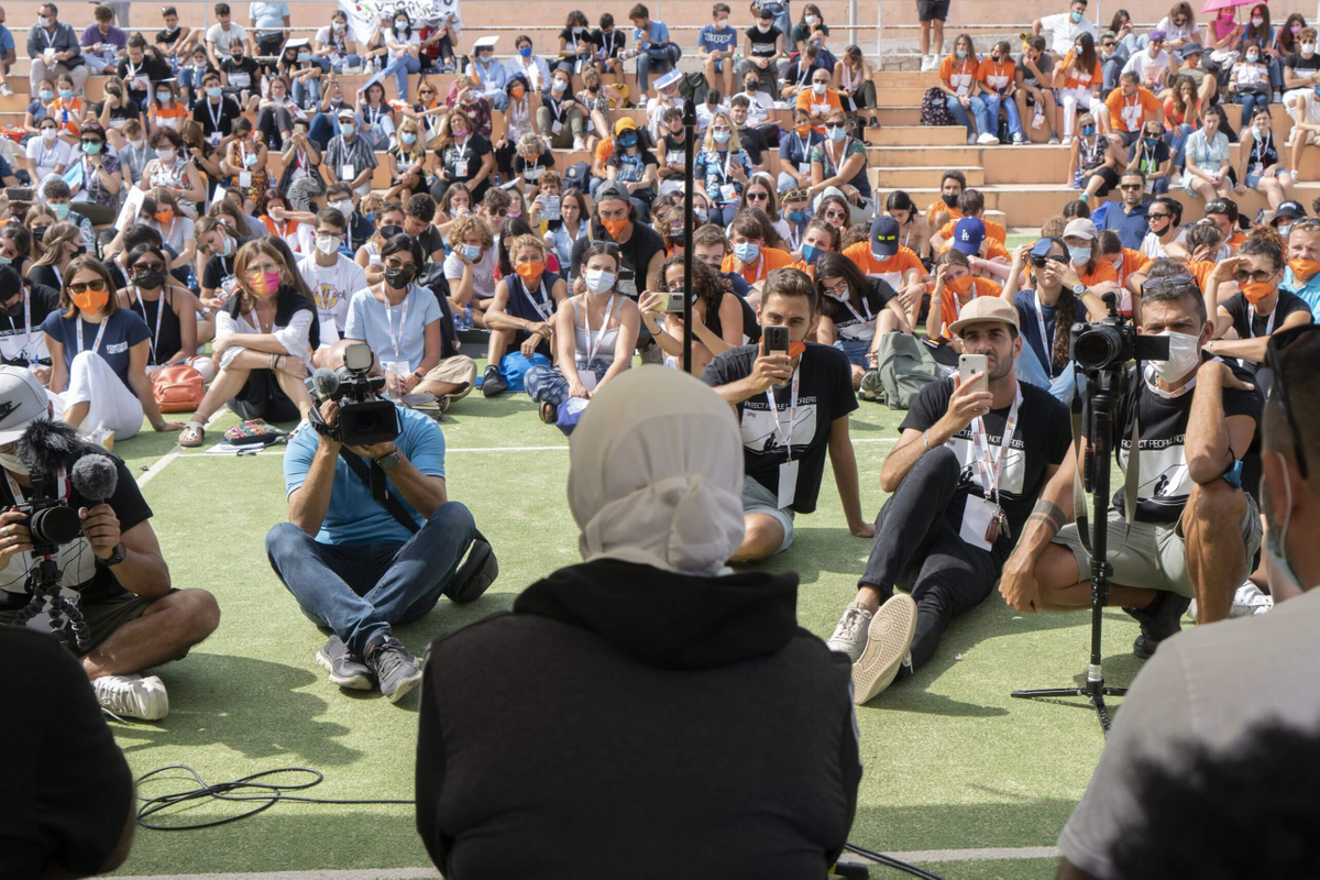 Un centinaio di persone che ascoltano le persone sul palco, con fotografi e video in primo piano